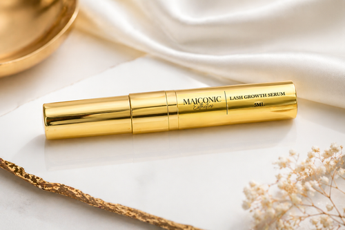 Gold MAICONIC lash growth serum bottle on a marble surface with decorative elements.